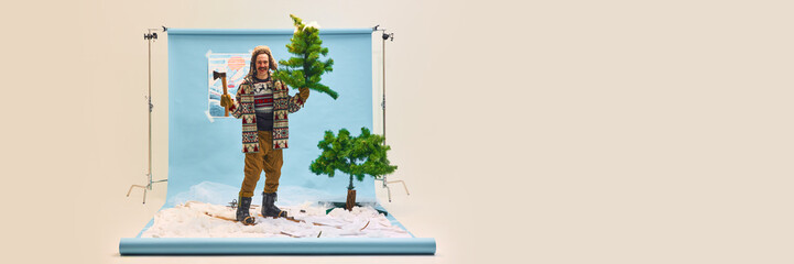 Man in winter attire with small artificial tree and axe, standing on fake snow and smiling. Concept...