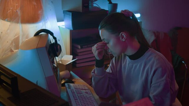 Tired young female developer taking off her glasses, rubbing nose bridge and eyebrows with closed eyes, then looking at vintage computer monitor