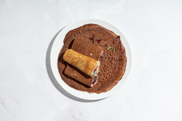 Close-up view of dark chocolate or buckwheat crepes, filled with greens and purple sprouts, served on a plate.