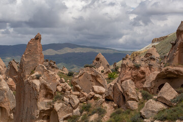 Ancient rock formations with carved dwellings rise over a green valley. A distant town sits at the foot of rolling hills under a cloudy sky, blending history, nature, and human presence in a breathtak