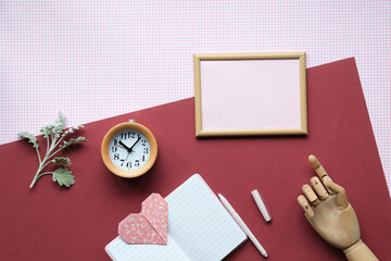 Flat lay of a workspace with a blank wooden frame, clock, notebook, paper heart, chalk, and mannequin hand on a red and pink background