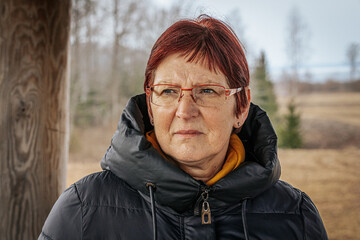 A portrait of an older woman with red hair and glasses, wearing a black winter coat. Her serious...