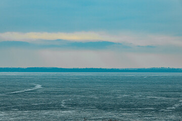 Obraz premium A frozen marshland with patches of dry grass and shrubs protruding through the icy surface. The distant shoreline fades into the cold, overcast sky, creating a serene winter scene.