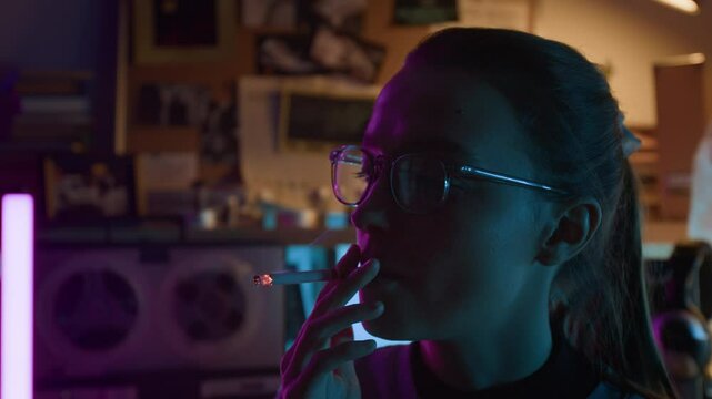 Close-up view of thoughtful young woman with glasses and hair in ponytail smoking cigarette in dimly lit room