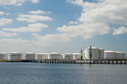 A wide, horizontal view of a waterfront industrial area, dominated by numerous large, white cylindrical storage tanks. 