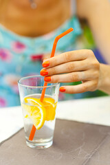 Close-up of a woman's hand holding a glass of lemon water decorated with a slice of lemon and a bright orange straw, showcasing vibrant summer vibes.