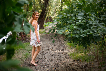 A joyful, barefoot woman in a floral summer dress enjoys a serene walk along a shaded forest path...