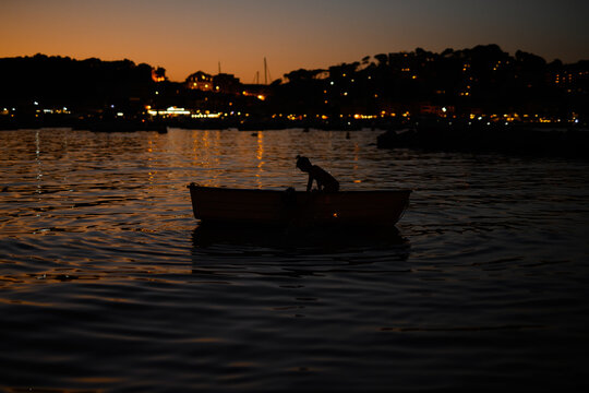 Silhouette of a Child in Boat at Dusk with Village Lights in Soller