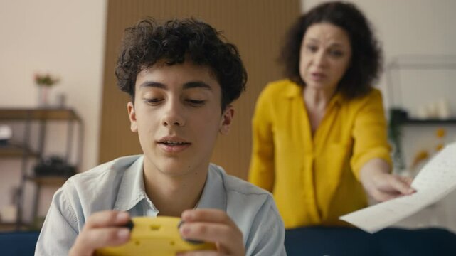Lazy teen son playing a video game and ignoring his mom showing bad test results