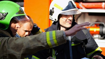 Male firefighters in protective uniforms discuss rescue plan looking at paper instructions near...
