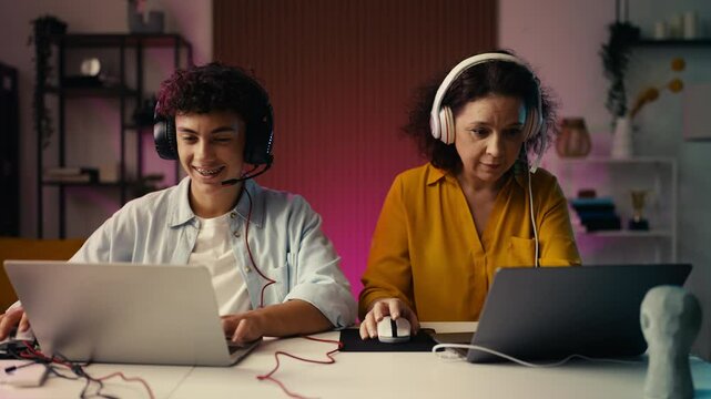 Happy mom and her teen son in headsets playing a game on laptops, bonding time