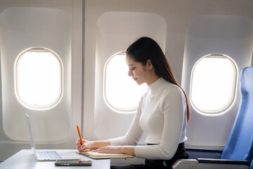 Fototapeta premium Aviation Business: An industrious woman is immersed in her work, jotting notes and managing her laptop on a plane, depicting modern business travel