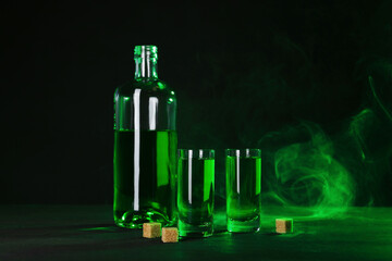 Absinthe in shot glasses, bottle and brown sugar on table against black background with smoke