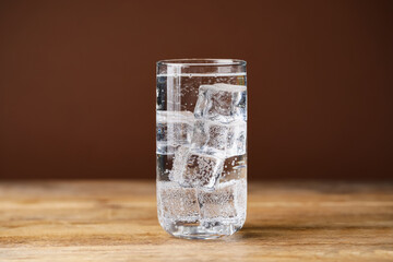 Refreshing soda water with ice cubes in glass on wooden table against brown background