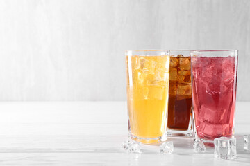 Soda water of different flavors in glasses with ice cubes on white wooden table, space for text