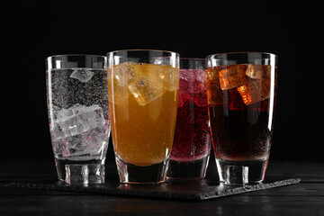Refreshing soda water of different flavors with ice cubes in glasses on black wooden table