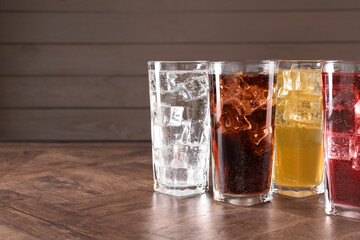 Refreshing soda water of different flavors with ice cubes in glasses on wooden table, space for text