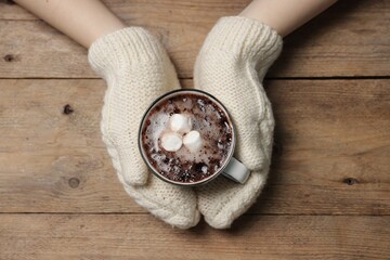 Woman holding mug of tasty hot chocolate with marshmallows at wooden table, top view