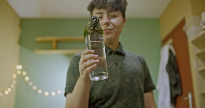 A girl offers a glass of water to her pet parrot in her bedroom, watching as the bird drinks and splashes happily, enjoying the cool refreshment.
