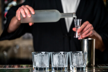 A bartender in a black outfit is pouring a clear drink into a shot glass while three glasses with ice sit on the counter, capturing a lively cocktail-making moment.