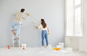 Young couple painting interior walls of their new apartment together, holding paint rollers and...