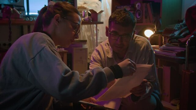 Tilt up view of two excited diverse young conspiracy theorists studying criminal profiles while sitting on carpet in dark messy room