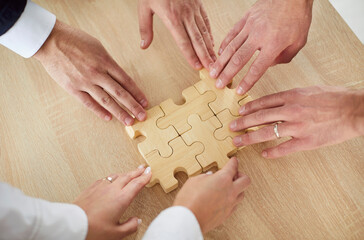 Top view cropped hands of a group of business people assembling wooden puzzle. Coworkers join jigsaw pieces in the office finding solution, working on project together. Support in teamwork concept.