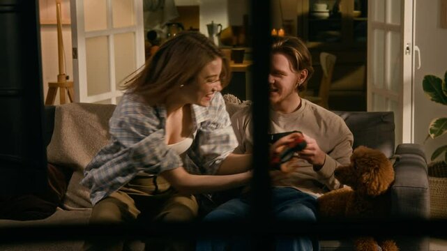 Medium full shot, seen through window from street, of young Caucasian man and woman sitting on couch at home, competing in video game using controllers, girl winning, hugging boyfriend and pet dog