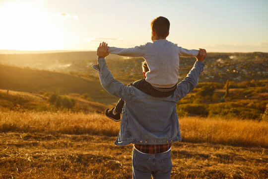 Back view portrait of young father and child boy sitting on shoulders holding hands and looking into the distance enjoying sunset and nature during a walk outdoors. Fathers day concept.