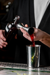 A bartender pours red liquid from a bottle into a metal jigger, preparing a cocktail. The focus is on the action and the sleek bar environment.