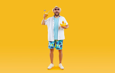 Happy male tourist enjoying cocktails on summer vacation. Cheerful funny bearded man in holiday shorts, shirt, sun hat and sunglasses holding drink and beach toy standing isolated on color background
