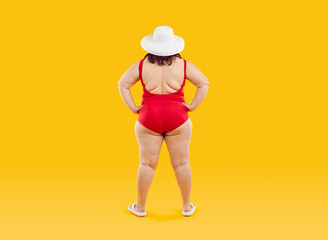 Full body photo of a funny fat overweight woman in red swimsuit and beach hat standing back with hands at sides isolated on studio yellow background. Summer holiday trip and vacation concept.