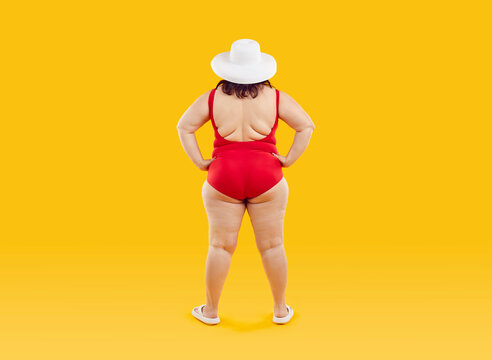 Full body photo of a funny fat overweight woman in red swimsuit and beach hat standing back with hands at sides isolated on studio yellow background. Summer holiday trip and vacation concept.
