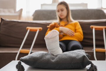 Broken leg. Young girl with broken leg cast sitting on sofa. Sad little kid recovering from broken...