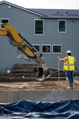 Fototapeta premium Construction worker in safety vest and hardhat working on new home foundation with excavator bucket scoop, job site for new housing development 