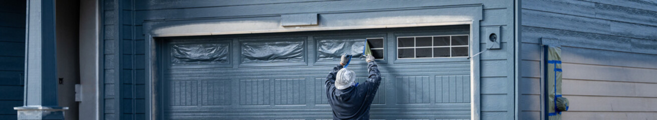 Professional painter in hard hat and air respirator removing brown masking paper from freshly painted garage door, job site for new housing development
