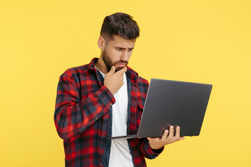 Thinking bearded hipster young man in plaid shirt using notebook or computer over yellow background