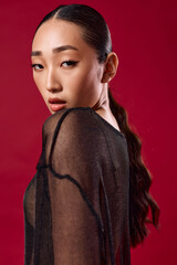 Fashion asian woman in sheer black top posing against red background with hands on hips and hair in ponytail