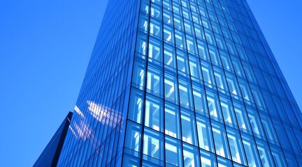 Obraz premium A modern skyscraper with a glass facade reflecting blue tones, showcasing striking architectural design and symmetry against a clear sky. 