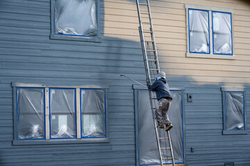 Professional painter in hard hat and air respirator on extension ladder with paint sprayer painting...