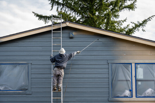 Professional painter in hard hat and air respirator on extension ladder with paint sprayer painting back exterior of house, job site for new housing development
