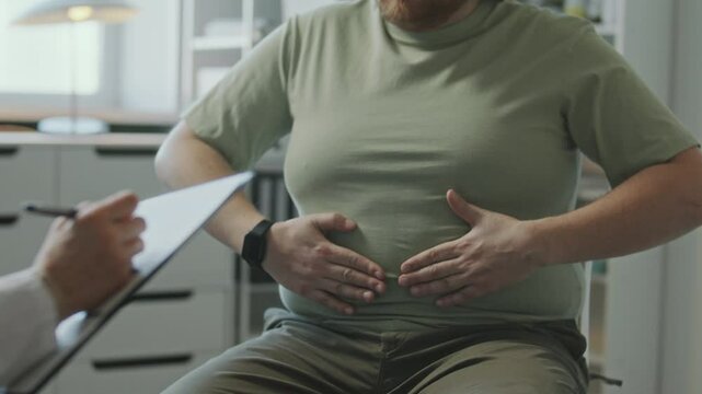 Male patient holding stomach and complaining of belly fat to female doctor taking notes on clipboard during weight loss consultation in clinic. Midsection shot