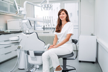 Portrait of sincerely smiling young dentist woman dressed white medical scrubs uniform sitting in...