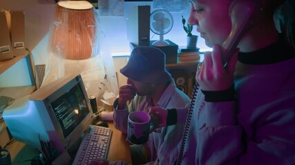 Positive girl holding mug pointing at vintage monitor with running programming code when speaking over corded telephone next to young developer with bucket hat who checking code lines for errors - Powered by Adobe