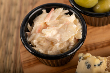 A black bowl of coleslaw sits on a wooden table. The bowl is filled with a creamy white dressing and is surrounded by a few pieces of cheese. The cheese is cut into small cubes
