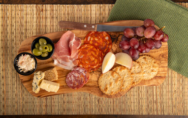 A wooden board top down shot with a variety of food items including crackers, cheese, grapes, olives, and meat