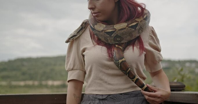 A woman with red hair stands with her pet snake around her neck, enjoying the day.