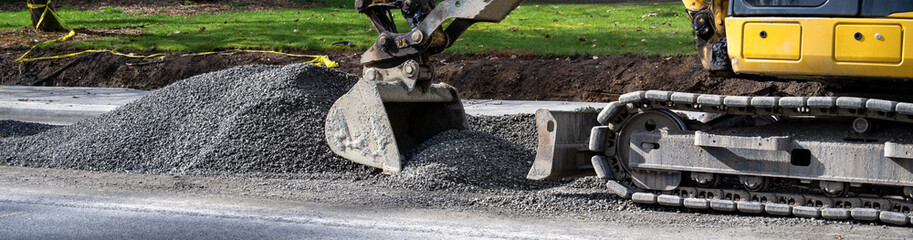 New public works sidewalk construction project between road and park, heavy equipment excavator with jawbone bucket and pile of gravel
