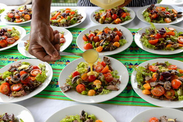 Hand pouring dressing over a gourmet salad on a white plate. Several similar salads on a table. The salad contains lettuce, red and yellow cherry tomatoes, olives and pieces of meat.Close-up