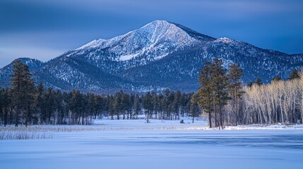 Majestic snow-capped mountain peak overlooking a frozen lake and pine forest at sunrise.